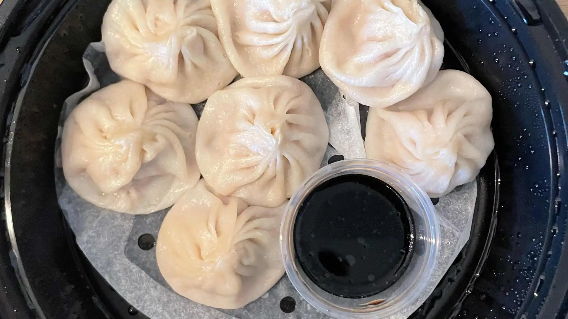 Soup dumplings on parchment in a black tray with a cup of dipping sauce at Juicy Dumpling Factory
